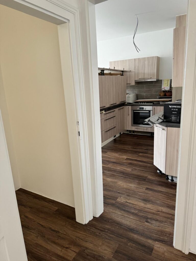 Moderní kuchyň propojená s chodbou, tmavá vinylová podlaha a nové kuchyňské skříňky v dekoru světlého dřeva.